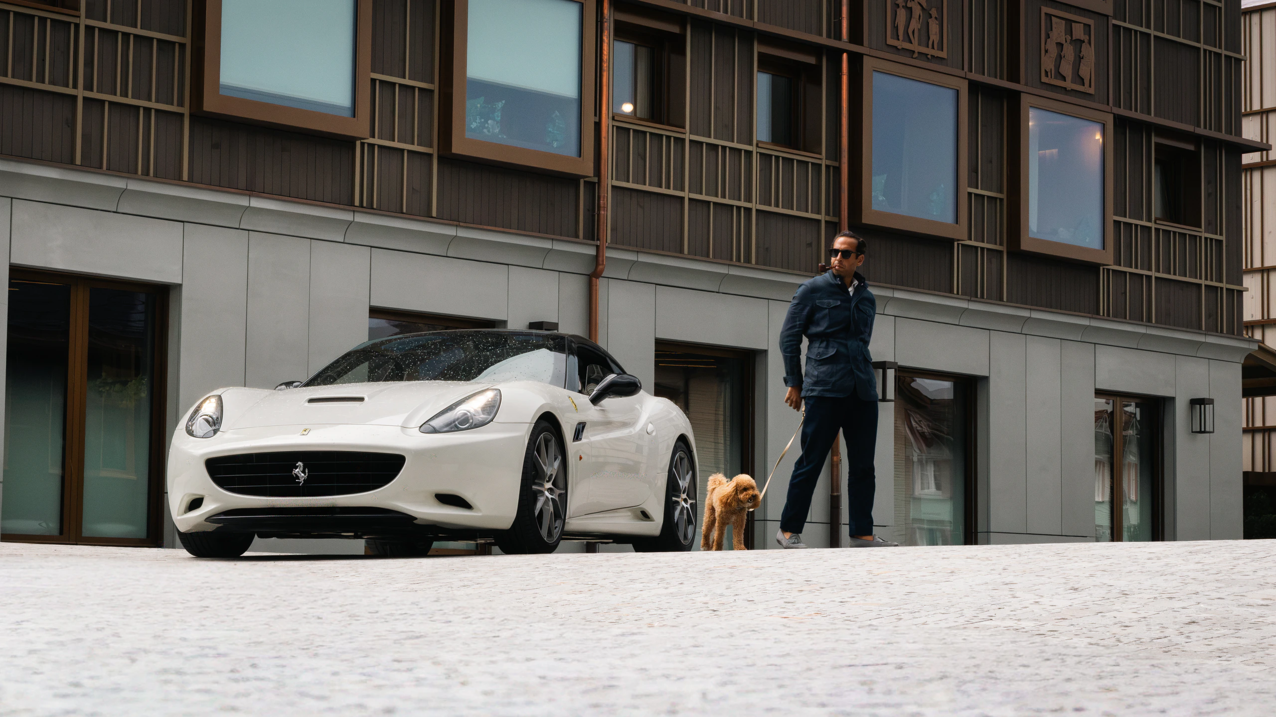 A white Ferrari California parked on cobbles outside the studio, a man walking a small dog past it, with the Shepherd Studios wordmark across the frame.