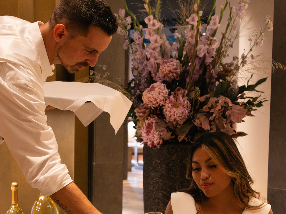 A waiter serving a seated guest at a table set with flowers and champagne